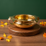 Brass Urli Bowl with side handles in a slight top-angle front view on a round wooden base with scattered marigold petals against a green backdrop