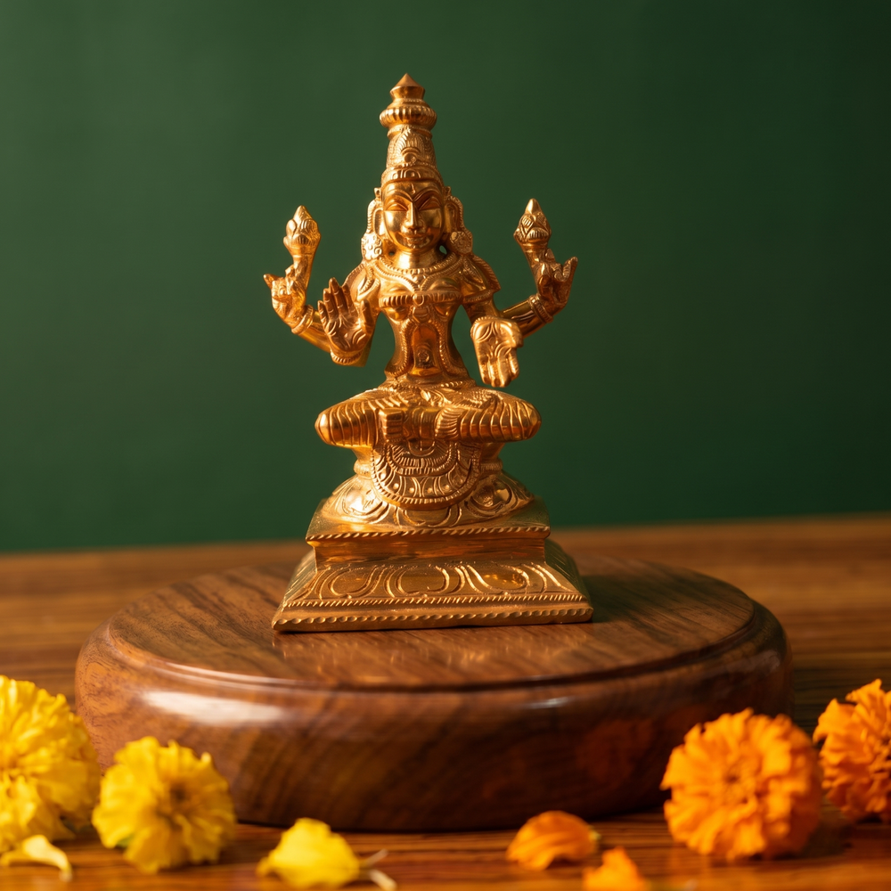 Lakshmi idol in front view, seated in lotus position with four arms on an engraved base with yellow and orange marigolds on a wooden plate.
