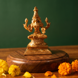 Lakshmi idol in front view, seated in lotus position with four arms on an engraved base with yellow and orange marigolds on a wooden plate.