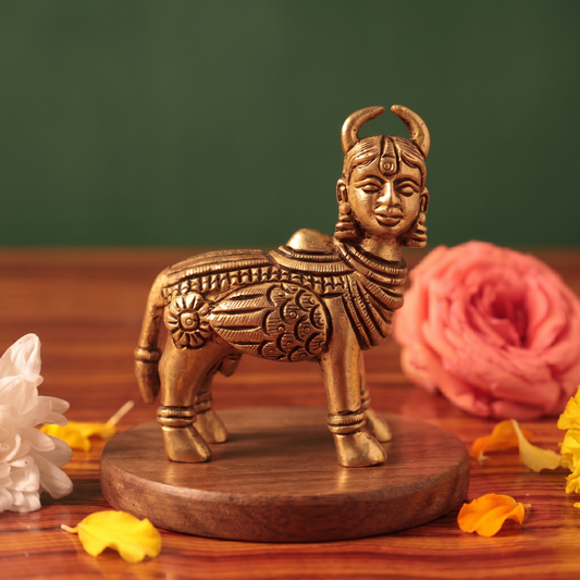 Brass Kamadhenu idol in straight front-facing standing view without calf, on a wooden platform with white and pink flowers, green backdrop.