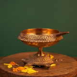 Aamai Vilakku Koorma Deepam with tortoise base in left-side view on a wooden base with scattered petals against a green backdrop