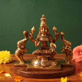 Brass Gaja Lakshmi idol in straight front-facing seated pose with two elephants on sides, on a wooden platform with flowers, green backdrop.