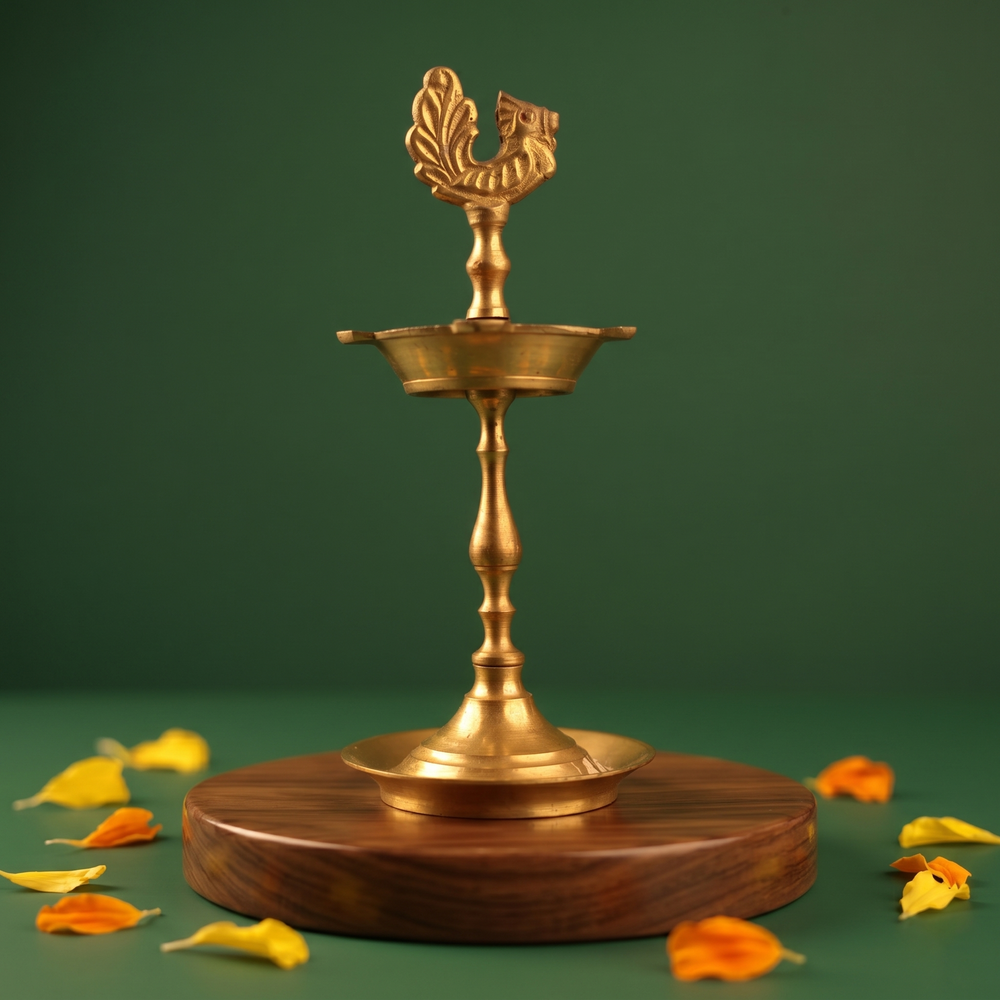 Karaikudi Vilakku with left-facing peacock on top in straight front view on a wooden base with colourful petals against a green backdrop