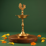 Karaikudi Vilakku with left-facing peacock on top in straight front view on a wooden base with colourful petals against a green backdrop