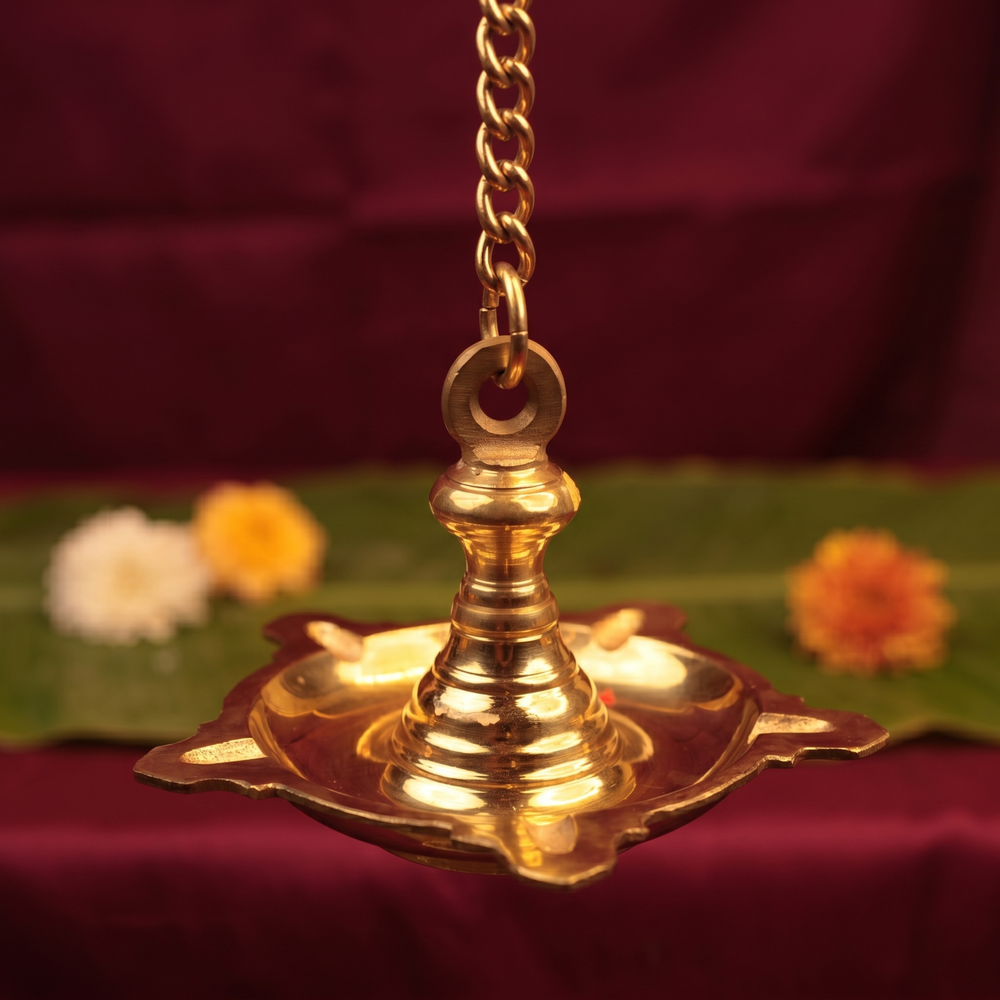 Thookku Vilakku hanging lamp with chain and scalloped base in close-up top-angle view with flowers on a banana leaf against a maroon backdrop