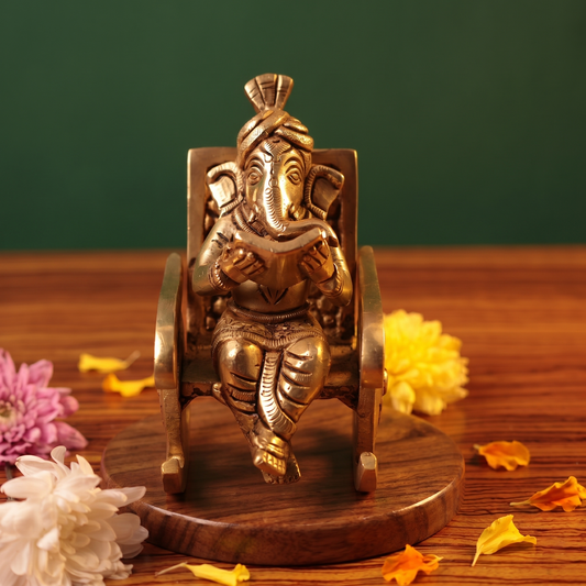 Brass Ganesh idol in straight front-facing pose, seated on a rocking chair reading a book, on a wooden platform with flowers, green backdrop.