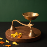 Brass Dhoopakal in left side angle with bell handle and deep bowl with scattered yellow and orange petals on wooden plate against dark background.