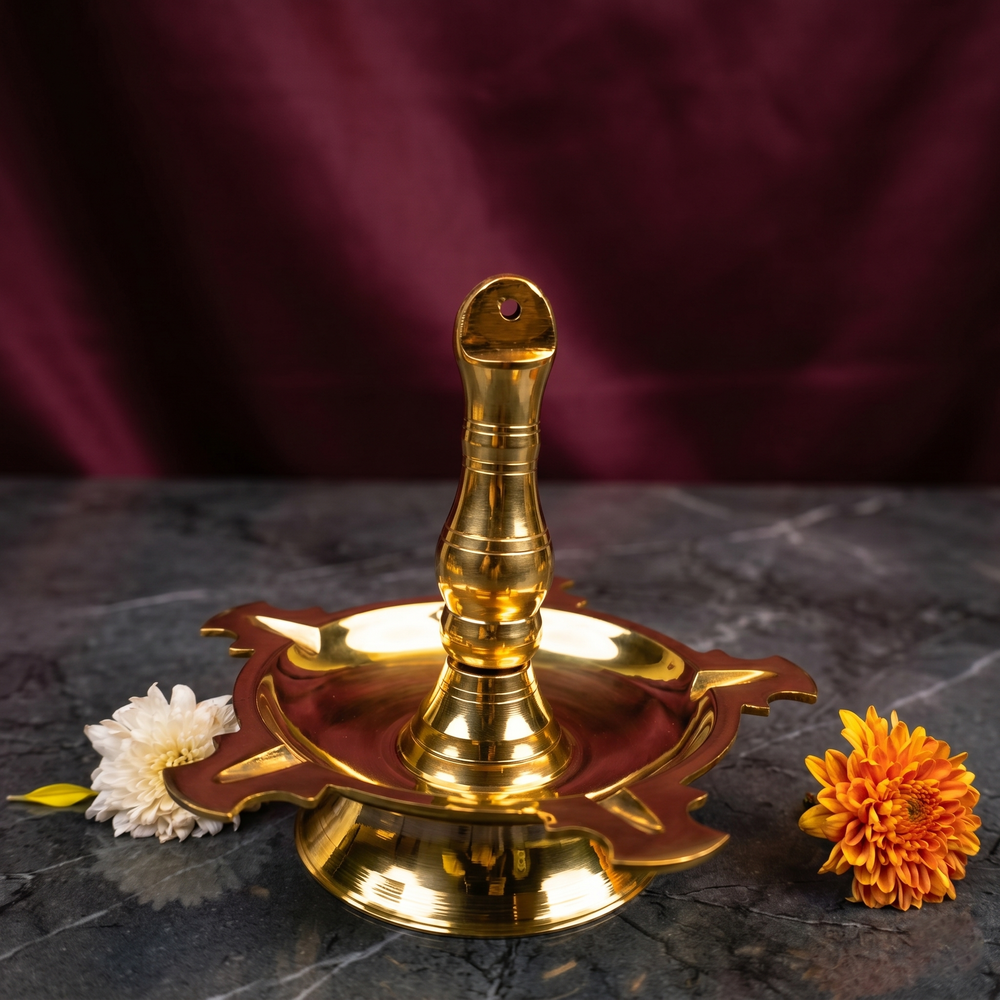 Thookku Vilakku with a scalloped base in a slight top-angle front view on a marble surface with white and orange flowers against a maroon backdrop