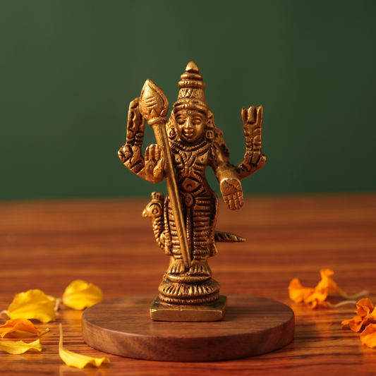Brass Murugan idol in straight front-facing standing pose holding vel with peacock at feet, on wooden platform with marigold petals, green backdrop.