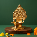 Kamakshi Deepam Vilakku with goddess on lotus in a slight right-angle view on a wooden base with marigolds against a green backdrop