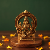 Brass Lakshmi idol in straight front-facing seated pose within decorative arch, on wooden platform with marigold petals, green backdrop.
