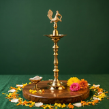 Annam Vilakku with left-facing swan on top in front view on wooden base with small diya and colourful flowers and petals against green backdrop