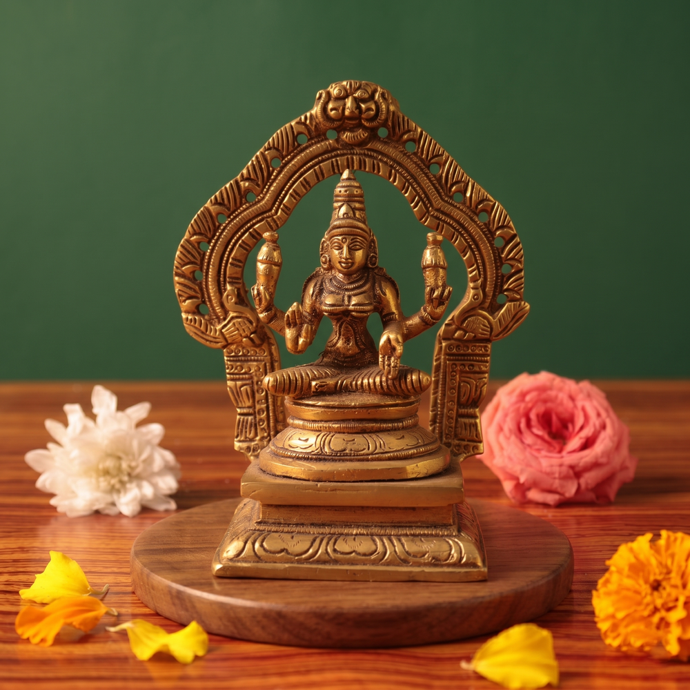 Brass Lakshmi idol in straight front-facing seated pose within an ornate arch, on a wooden platform with white and pink flowers, green backdrop.