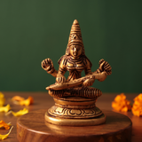 Brass Saraswathi idol in straight front-facing seated pose playing veena, on a wooden platform with marigold petals, green backdrop.