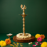 Annam Vilakku with left-facing swan on top in front view on wooden base with jasmine garland and colourful flowers against green backdrop