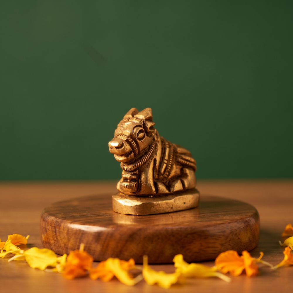 Brass Nandi idol in a slight right-angle resting pose on a wooden platform with scattered marigold petals, green backdrop.