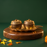 Two brass Kumkum Sindoor Boxes with lids on a scalloped brass plate in straight front view on a wooden base against a green backdrop