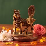 Brass Kamadhenu idol in a slight left-angle view with calf beneath, on a wooden platform with white and pink flowers, green backdrop.