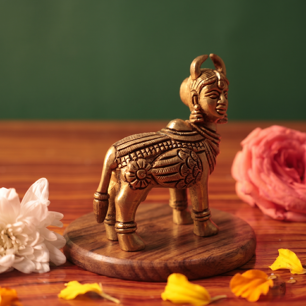 Brass Kamadhenu idol in left side-angle standing view without calf, on a wooden platform with white and pink flowers, green backdrop.