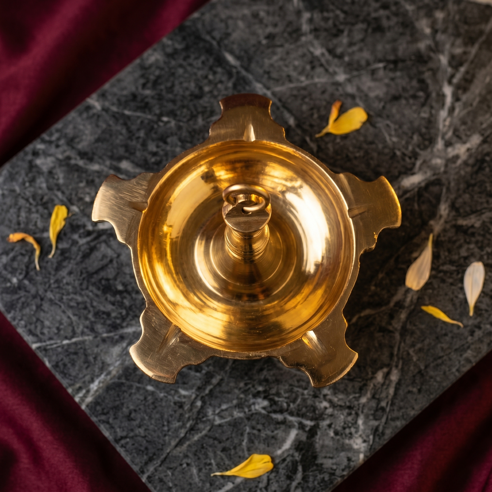 Thookku Vilakku with a scalloped base in top-down view on a marble surface with scattered petals against a maroon backdrop