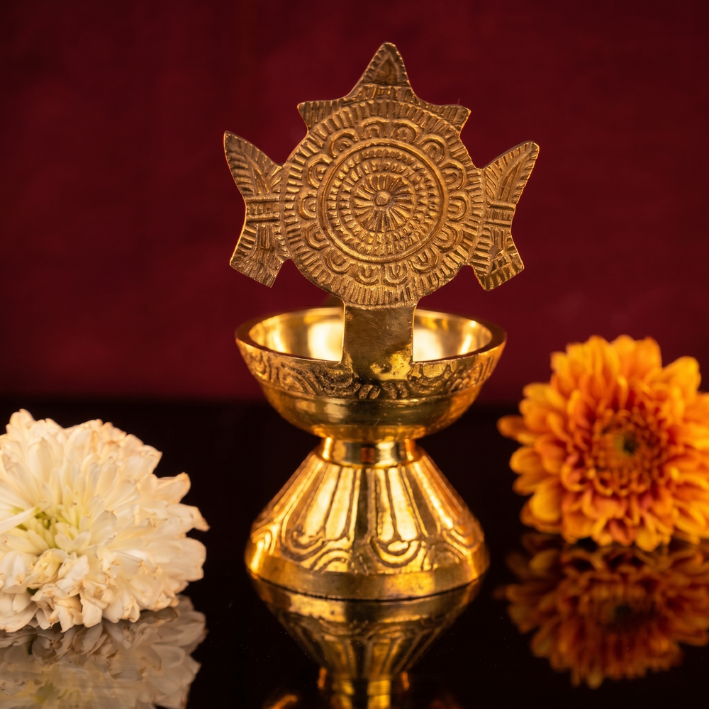 Sangu Chakaram Vilakku with engraved chakra in front view with white and orange flowers on reflective surface against a maroon backdrop