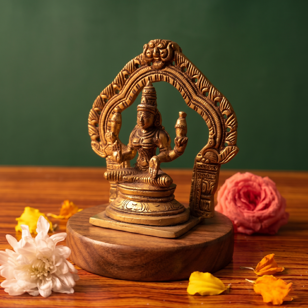 Brass Lakshmi idol in a slight right-angle seated view within an ornate arch, on a wooden platform with white and pink flowers, green backdrop.