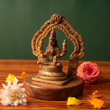 Brass Lakshmi idol in a slight right-angle seated view within an ornate arch, on a wooden platform with white and pink flowers, green backdrop.