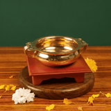 Brass Urli Bowl with side handles in front view on a maroon wooden stand with white and yellow flowers and petals against a green backdrop