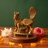 Brass Kamadhenu idol in a slight right-angle view with calf beneath, on a wooden platform with white and pink flowers, green backdrop.