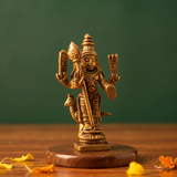 Brass Murugan idol in a slight left-angle standing view holding vel with peacock at side, on a wooden platform with petals, green backdrop.