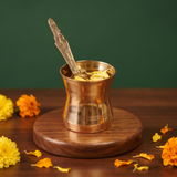 Brass Panchapatra and Achmani Set with engraved spoon and petals inside in front view on a wooden base with marigolds against a green backdrop