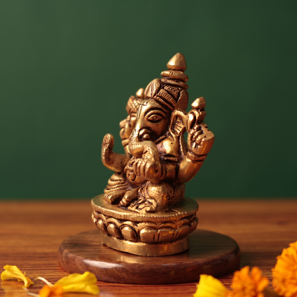 Brass Ganesh idol in left side-angle seated pose on lotus base, on wooden platform with marigold petals, green backdrop.