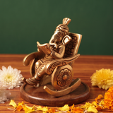 Brass Ganesh idol in left side-angle view, seated on a rocking chair reading a book, on a wooden platform with scattered flowers, green backdrop.