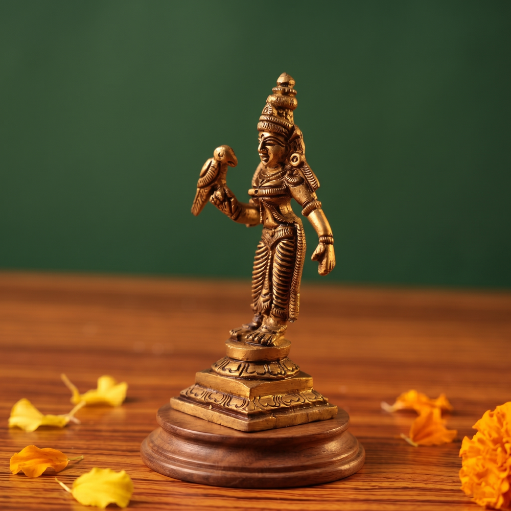 Brass Meenakshi idol in a slight right-angle standing view holding a parrot, on a wooden platform with scattered marigold petals, green backdrop.