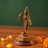 Brass Meenakshi idol in a slight right-angle standing view holding a parrot, on a wooden platform with scattered marigold petals, green backdrop.