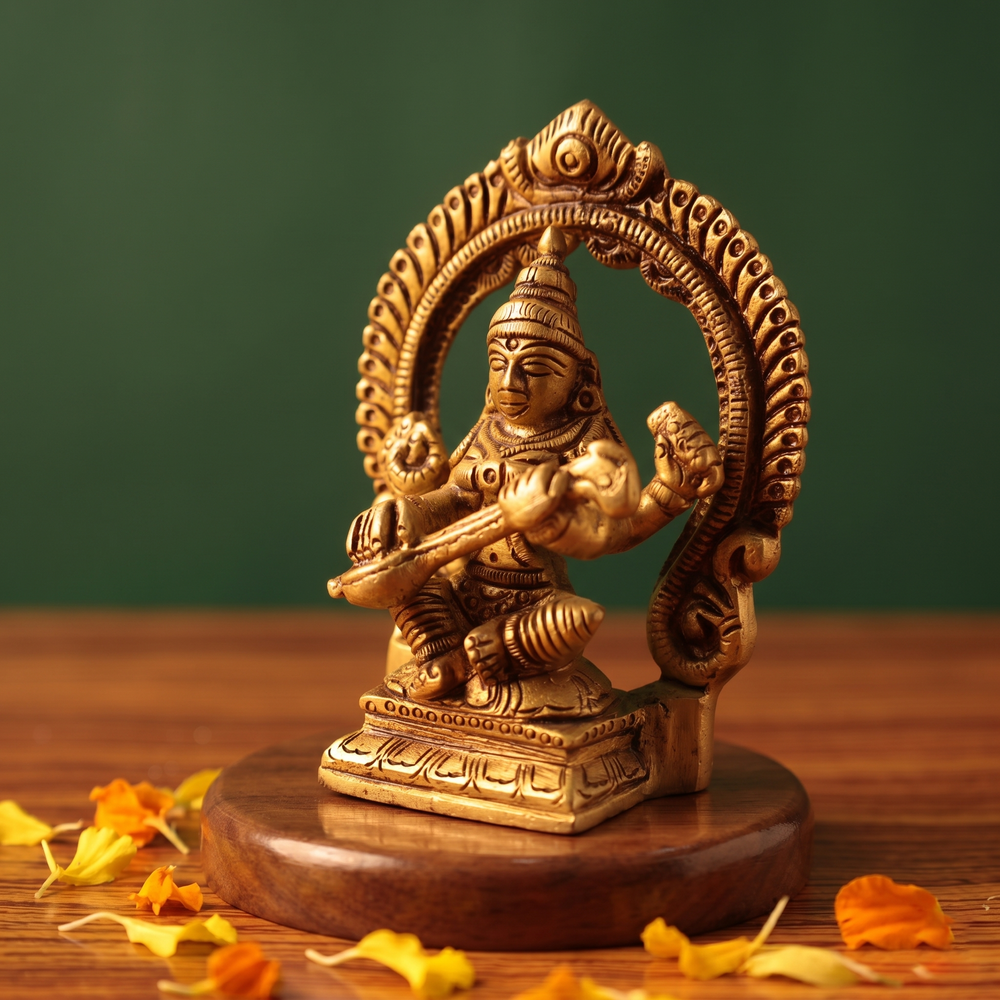 Brass Saraswathi idol in a slight right-angle seated pose playing veena within a decorative arch, on a wooden platform, green backdrop.