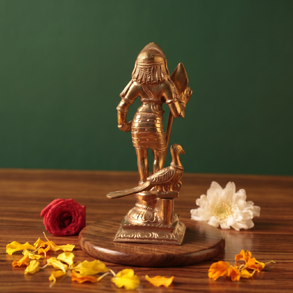 Bala Murugan with Peacock in back view holding vel with peacock at base with red and white flowers and scattered petals on a wooden plate.