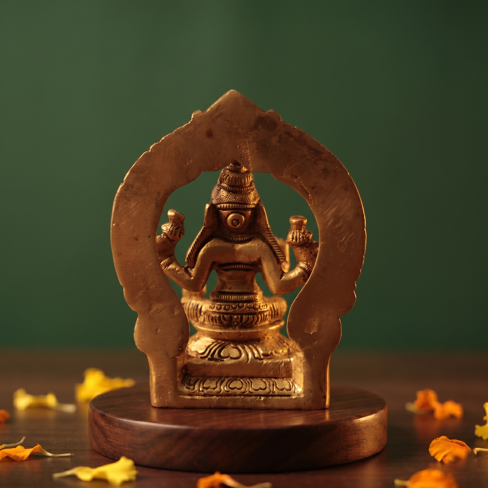 Brass Lakshmi idol back view seated within decorative arch, on wooden platform with scattered marigold petals, green backdrop.