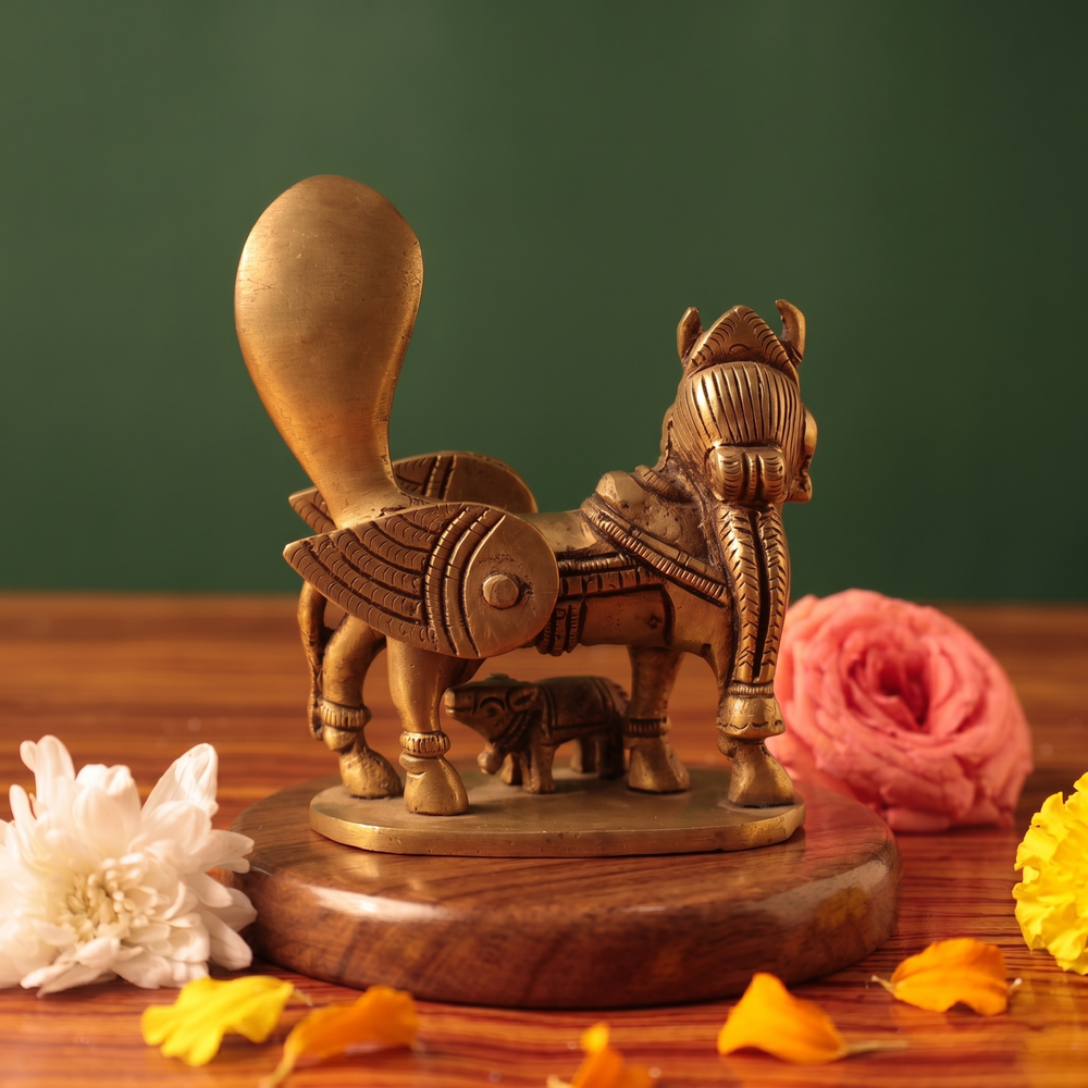 Brass Kamadhenu idol back view with calf beneath, on a wooden platform with white and pink flowers, green backdrop.