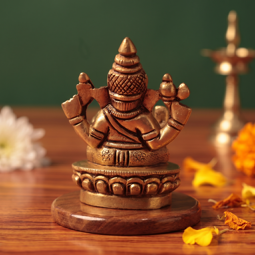 Brass Ganesh idol back view, seated on lotus base, on wooden platform with flowers and diya, green backdrop.