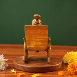 Brass Ganesh idol back view, seated on a rocking chair, on a wooden platform with white and yellow flowers, green backdrop.