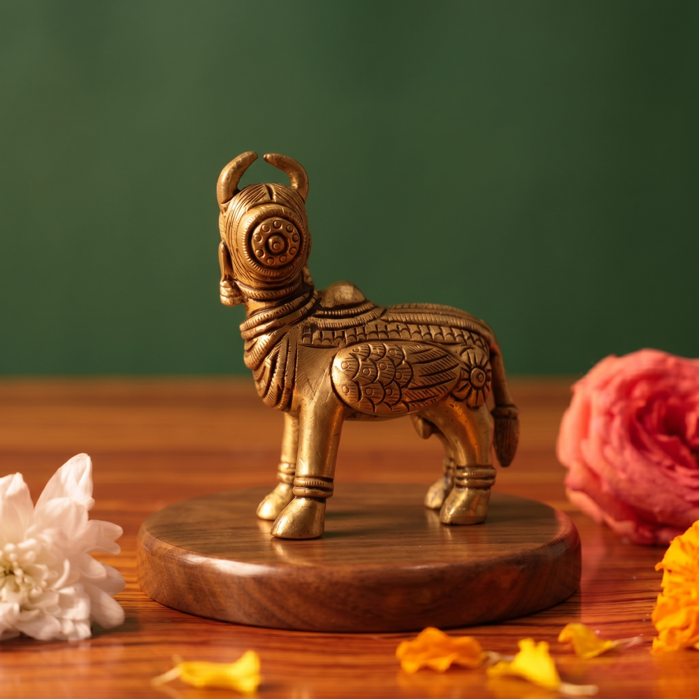 Brass Kamadhenu idol back view standing without calf, on a wooden platform with white and pink flowers, green backdrop.