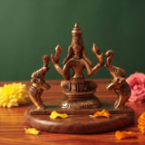 Brass Gaja Lakshmi idol back view, seated and flanked by two elephants, on a wooden platform with scattered petals, green backdrop.