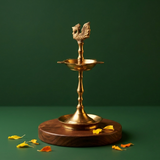 Karaikudi Vilakku with right-facing peacock on top in straight front view on a wooden base with scattered petals against a green backdrop