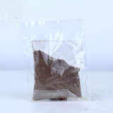 Thirtha parimalam powder in a clear sealed plastic packet showing dark brown aromatic powder in straight front view against white backdrop.