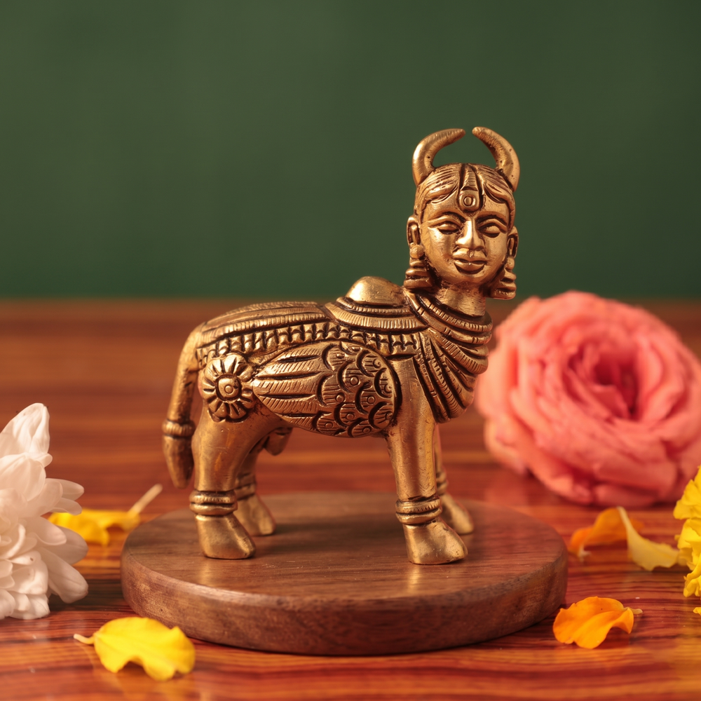 Brass Kamadhenu idol in straight front-facing standing view without calf, on a wooden platform with white and pink flowers, green backdrop.

