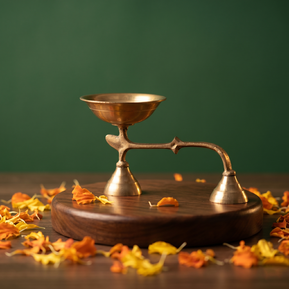 Brass Dhoopakal in right side angle with bell handle and deep bowl with scattered yellow and orange petals on wooden plate against green background.