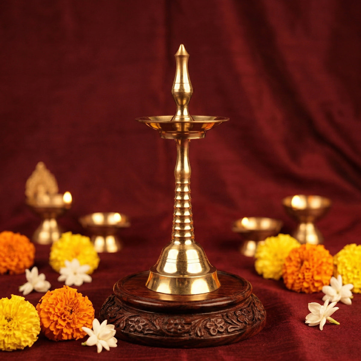 Nilavilakku Kerala floor lamp in straight front view on carved wooden base with marigold and jasmine flowers against a maroon backdrop