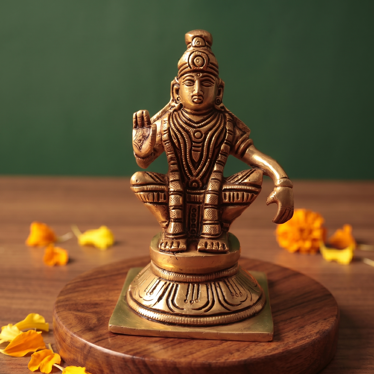 Brass Ayyappan statue in straight front-facing seated pose, raising right hand in blessing, on wooden platform with marigolds, green backdrop.
