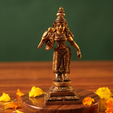 Brass Meenakshi idol in a straight front-facing standing pose holding a parrot, on a wooden platform with marigold petals, green backdrop.
